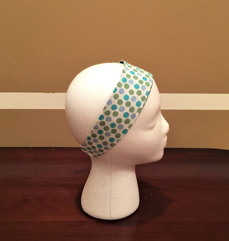 May include: A fabric headband featuring a pattern of blue and green polka dots on a white background. The headband is displayed on a white mannequin head. The headband is approximately 2.5 cm wide.