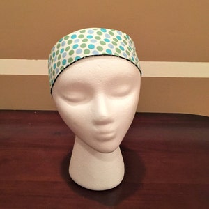 May include: A white headband patterned with blue and green polka dots. The headband is displayed on a white mannequin head. The edges of the headband are trimmed with black stitching. A hair accessory.