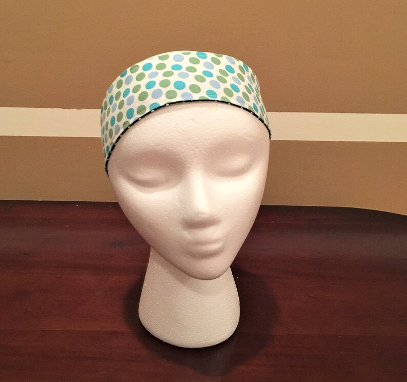 May include: A white headband patterned with blue and green polka dots. The headband is displayed on a white mannequin head. The edges of the headband are trimmed with black stitching. A hair accessory.