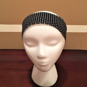 May include: A black headband with a white polka dot pattern is shown on a mannequin head. The fabric headband has small white dots on a black background. The accessory is designed to be worn around the head, and is suitable for various activities.