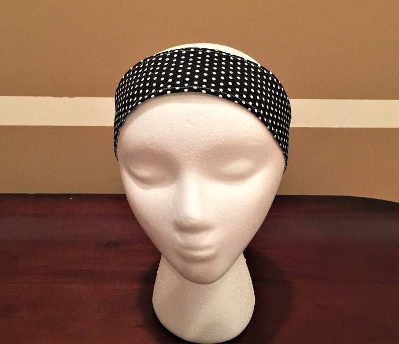 May include: A black headband with a white polka dot pattern is shown on a mannequin head. The fabric headband has small white dots on a black background. The accessory is designed to be worn around the head, and is suitable for various activities.
