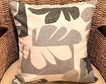 May include: Square decorative pillow with a neutral color palette. The pillow features a large-scale leaf pattern in shades of gray, beige, and white. The pillow is placed on a wicker chair, suggesting a natural or coastal theme.