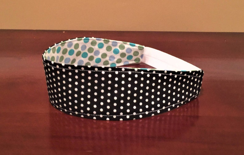 May include: A black and white polka dot headband with a white elastic band. The interior lining features a pattern of teal and green polka dots on a white background. This accessory is designed for comfort and style.