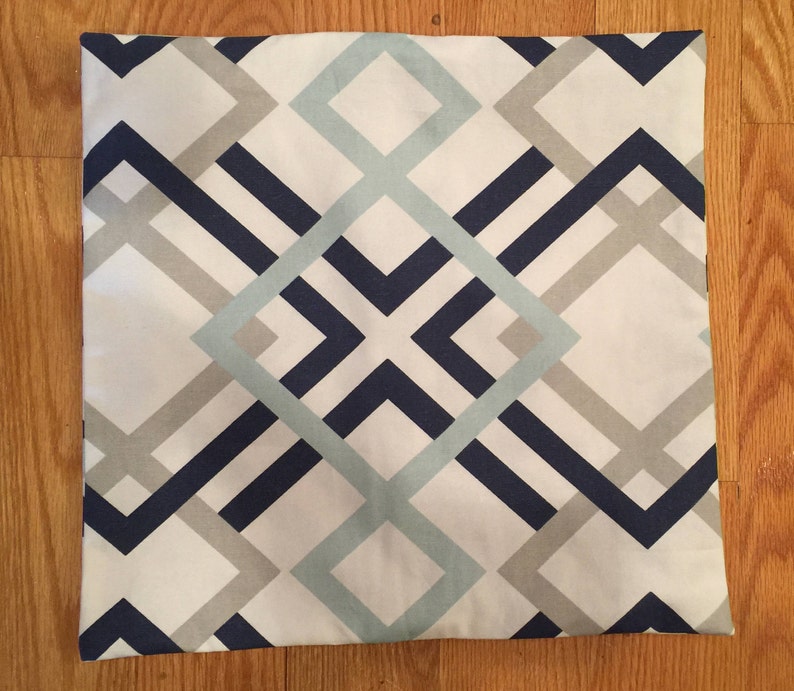 May include: Square pillow with a geometric pattern. The design features intersecting lines and shapes in navy blue, light blue, and gray against a white background. The pillow is square and appears to be made of fabric.