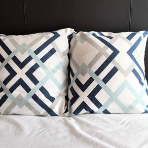 May include: Two square decorative pillows with a geometric pattern. The pattern features intersecting lines in navy blue, light blue, and gray on a white background. The pillows are on a white bed.