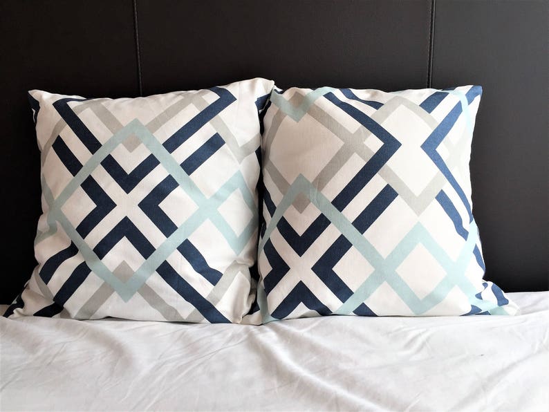 May include: Two square decorative pillows with a geometric pattern. The pattern features intersecting lines in navy blue, light blue, and gray on a white background. The pillows are on a white bed.