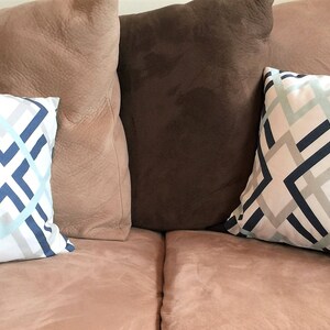 May include: Two decorative throw pillows with a geometric pattern in blue, light blue, and gray on a white background. The pillows are placed on a pinkish-brown sofa, with other solid-colored pillows in the background.