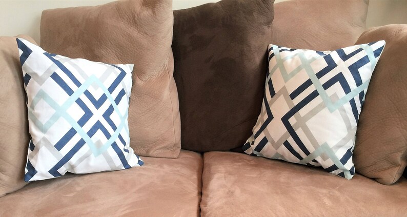 May include: Two decorative throw pillows with a geometric pattern in blue, light blue, and gray on a white background. The pillows are placed on a pinkish-brown sofa, with other solid-colored pillows in the background.