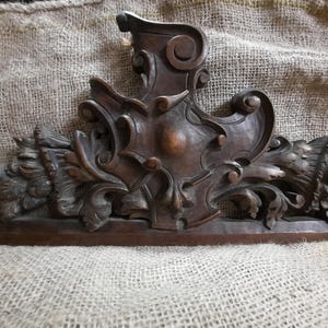 Può includere: Un frontone architettonico in legno marrone scuro caratterizzato da intricati intagli a mano a volute, motivi a foglie d'acanto e un emblema centrale a forma di scudo. L'elemento decorativo presenta una base piatta e intagli simmetrici.