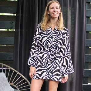 May include: A black and white zebra print dress with long sleeves and a tie waist. The dress is knee-length and worn by a person standing outdoors. The background includes a black curtain and a wooden fence.