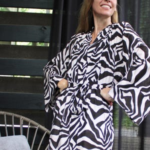 May include: A black and white zebra print kimono-style robe with wide sleeves and a tie waist. The robe is knee-length and made of a lightweight fabric. The background includes a black curtain and a wooden fence.
