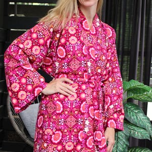 May include: A long, floral-patterned robe in shades of pink, fuchsia, and orange. The robe features a tie waist and long sleeves. The design includes a repeating pattern of stylized flowers and geometric shapes. The robe is made of a soft, flowing fabric.