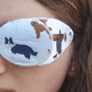 May include: A white eye patch with a brown and black dog print. The eye patch is made of soft fabric and has a white stitched border.