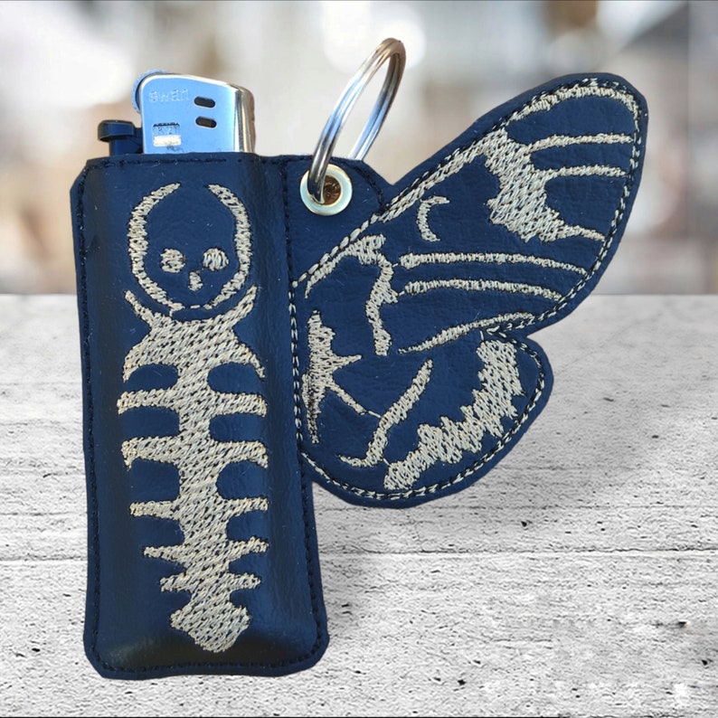 May include: A black leather lighter case with a skeleton and butterfly design. The case has a silver metal ring and a zipper closure.