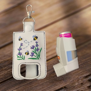 May include: White leather keychain holder with an embroidered design of bees and lavender flowers. The holder has a clip and a loop for attaching to a bag or belt. A white inhaler is also pictured.