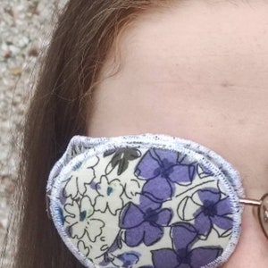 May include: An eye patch with a floral pattern in shades of purple and white. The patch is oval-shaped and has a white trim. The design features large purple flowers and smaller white flowers on a cream background.