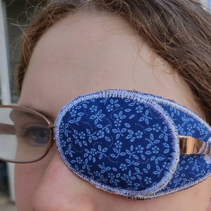 May include: A blue eye patch with a white floral pattern. The patch is made of fabric and has a white border. It is being worn over a person's right eye.