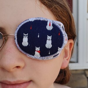 May include: A close-up of a person wearing a blue eye patch with a white cat pattern. The eye patch has a white border and is secured with an elastic strap.