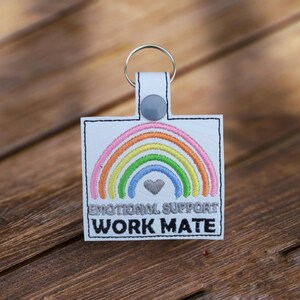 May include: White leather keychain with a rainbow and a heart embroidered on it. The text "Emotional Support Work Mate" is also embroidered on the keychain.