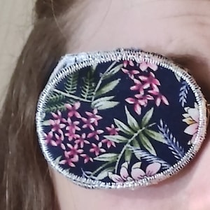 May include: A round eye patch with a floral print. The fabric is dark blue with pink and green flowers and leaves. The edge of the patch is trimmed with silver.