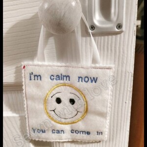 May include: White door with a white doorknob and a white fabric sign hanging from it. The sign says "I'm calm now you can come in" and has a yellow smiley face with black eyes.
