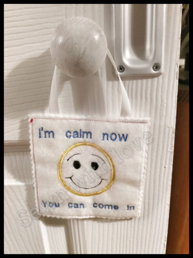 May include: White door with a white doorknob and a white fabric sign hanging from it. The sign says "I'm calm now you can come in" and has a yellow smiley face with black eyes.