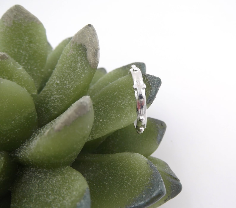 May include: A silver rosary ring with small beads and a cross design. The ring is displayed on a green succulent plant.