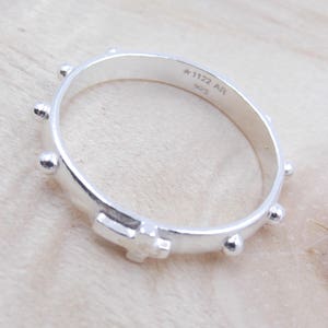 May include: Silver rosary ring with a cross and small beads. The ring is engraved with the text "A-1122 AR 925".
