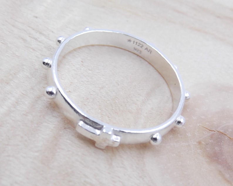 May include: Silver rosary ring with a cross and small beads. The ring is engraved with the text "A-1122 AR 925".