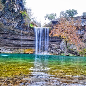 May include: A waterfall cascades into a clear, turquoise pool surrounded by rocky cliffs. The water is a vibrant green and blue, and the rocks are a mix of gray and brown. Lush greenery surrounds the pool, adding to the natural beauty of the scene.