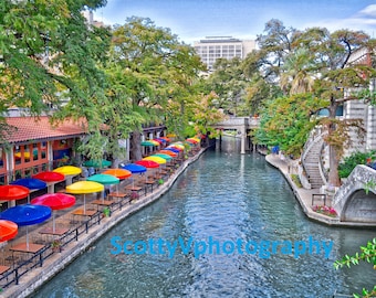 May include: A view of a river with colorful umbrellas lining the edge of the river. The umbrellas are over tables and chairs, suggesting an outdoor dining area. There are buildings and trees on the other side of the river.