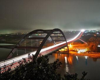 May include: A long exposure photograph of a bridge lit up at night with cars driving across it. The bridge is a large arch bridge with a concrete deck and steel supports. The bridge is lit up with white lights, and the cars are leaving long streaks of light as they drive across the bridge. The bridge is over a body of water, and the water is reflecting the lights of the bridge. The bridge is surrounded by trees and bushes, and the sky is dark and cloudy.