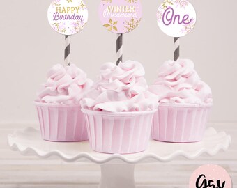 Puede incluir: Tres cupcakes rosas con glaseado blanco y adornos de cupcakes con temática de copos de nieve dorados y morados que dicen "Feliz cumpleaños", "Invierno mágico" y "Uno".
