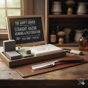 Puede incluir: Una navaja recta con mango de madera descansa sobre una almohadilla de cuero marrón. Una pequeña pizarra muestra "THE HAPPY SHAVER STRAIGHT RAZOR HONING & RESTORATION ETSY SHOP - Find Us!" También se ven varias piedras de afilar y una bandeja de madera.