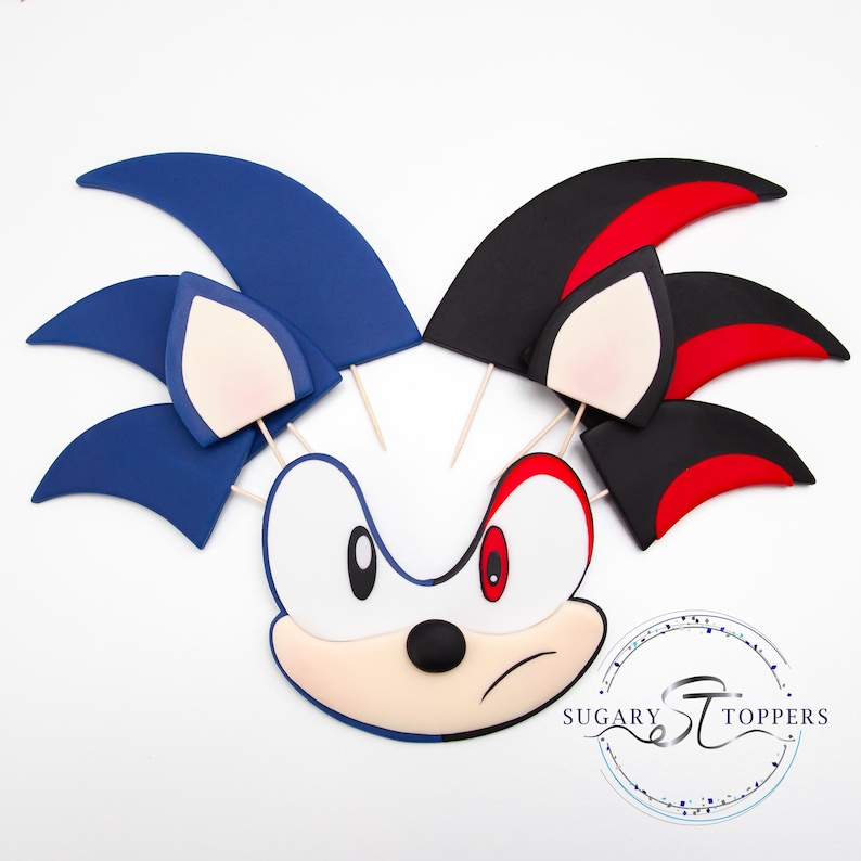 May include: Cake toppers featuring the faces of Sonic and Shadow the Hedgehog. The Sonic topper is blue, white, and tan, while the Shadow topper is black, red, and tan. Both have wooden sticks attached for easy placement on a cake. The Sugary Toppers logo is in the bottom right.