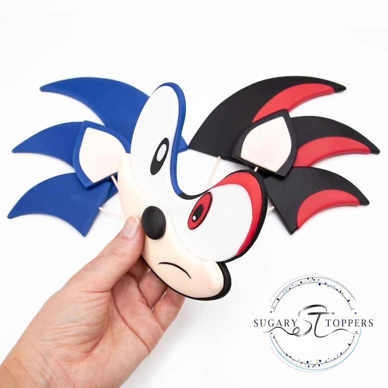 May include: A blue, white, and black Sonic the Hedgehog cake topper. The topper features the character's face and quills. The face is white with a black nose and eyes, and red accents. The quills are blue, black, and red.
