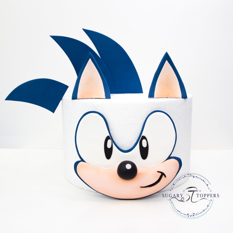 May include: A white cake decorated with a cartoon character's face, featuring blue spikes, large eyes, and a smiling mouth. The character's face is outlined in blue, with a peach-colored mouth and ears. The cake is a fun, themed dessert.