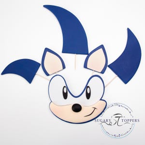 May include: A cartoon character face with blue spikes and ears, a white face, and a black nose and eyes. The character's face is smiling. The image includes the text "Sugary Toppers" in a circular design.