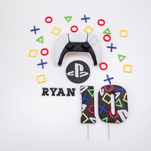 May include: A white and black game controller with a black PlayStation logo, the name "RYAN", and the number "10" made of black icing with colorful shapes. Red, blue, green, and yellow shapes are scattered around the controller.