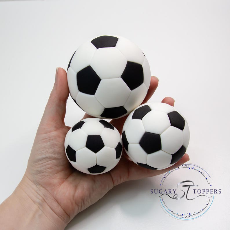 Pu&ograve; includere: Tre decorazioni sferiche a forma di pallone da calcio con un classico motivo a pentagoni bianchi e neri. Il set include un pallone grande e due pi&ugrave; piccoli, tutti con una finitura liscia. Il testo Sugary Toppers &egrave; visibile nell'angolo.