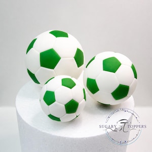 Pu&ograve; includere: Tre decorazioni sferiche a forma di pallone da calcio con un classico motivo a pentagoni bianchi e verdi. Il set comprende una palla grande, una media e una piccola, disposte insieme. Il testo Sugary Toppers &egrave; visibile.