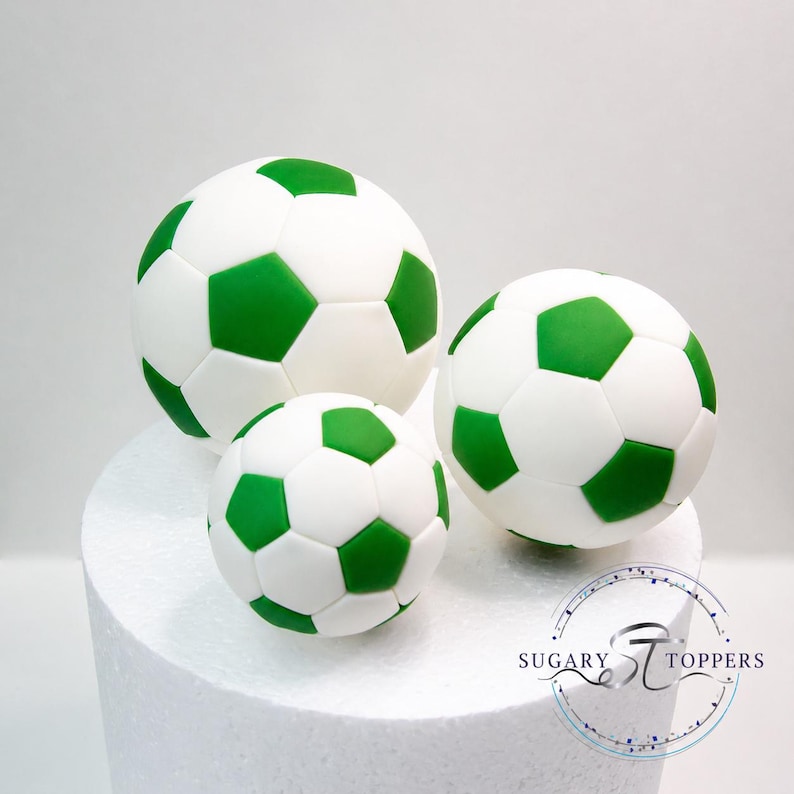 Pu&ograve; includere: Tre decorazioni sferiche a forma di pallone da calcio con un classico motivo a pentagoni bianchi e verdi. Il set comprende una palla grande, una media e una piccola, disposte insieme. Il testo Sugary Toppers &egrave; visibile.