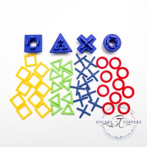 May include: A collection of colorful geometric shapes, including squares, triangles, crosses, and circles. The shapes are in blue, yellow, green, and red. The image includes the text "SUGARY TOPPERS".