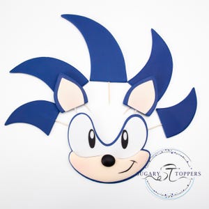 May include: A blue and white Sonic the Hedgehog cake topper. The topper features the character's face with large eyes, a black nose, and a smiling mouth. The character's blue quills are attached to the head with wooden sticks.