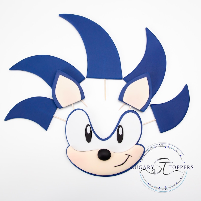 May include: A blue and white Sonic the Hedgehog cake topper. The topper features the character's face with large eyes, a black nose, and a smiling mouth. The character's blue quills are attached to the head with wooden sticks.