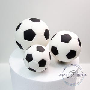 Pu&ograve; includere: Tre decorazioni per torte a forma di pallone da calcio di varie dimensioni, caratterizzate da un classico motivo a pentagoni ed esagoni bianchi e neri. Gli oggetti lisci e rotondi sono disposti insieme, con il logo SUGARY STOPPERS visibile.