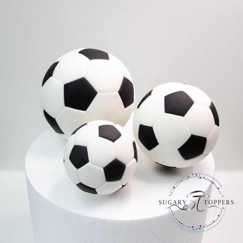 Pu&ograve; includere: Tre decorazioni per torte a forma di pallone da calcio di varie dimensioni, caratterizzate da un classico motivo a pentagoni ed esagoni bianchi e neri. Gli oggetti lisci e rotondi sono disposti insieme, con il logo SUGARY STOPPERS visibile.
