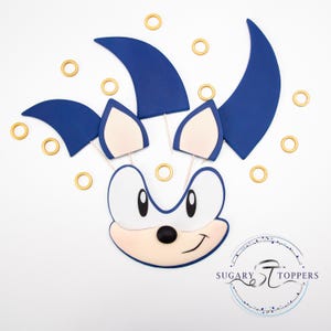 May include: A blue and white cartoon character face with blue spikes and gold rings. The character has a smiling expression and is likely a cake topper. The logo "Sugary Toppers" is in the bottom right corner.