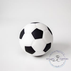 Pu&ograve; includere: Un topper per torta a forma di pallone da calcio sferico con il classico motivo a pentagoni ed esagoni bianchi e neri. La superficie presenta dettagli di cucitura visibili tra i pannelli. Il logo Sugary Toppers &egrave; stampato in basso a destra.