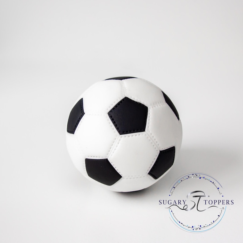 Pu&ograve; includere: Un topper per torta a forma di pallone da calcio sferico con il classico motivo a pentagoni ed esagoni bianchi e neri. La superficie presenta dettagli di cucitura visibili tra i pannelli. Il logo Sugary Toppers &egrave; stampato in basso a destra.